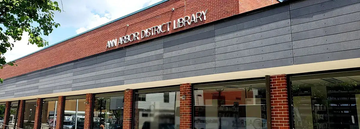 Exterior of the Ann Arbor District Library building, showing its red brick and dark paneled facade with an illuminated "Ann Arbor District Library" wall sign