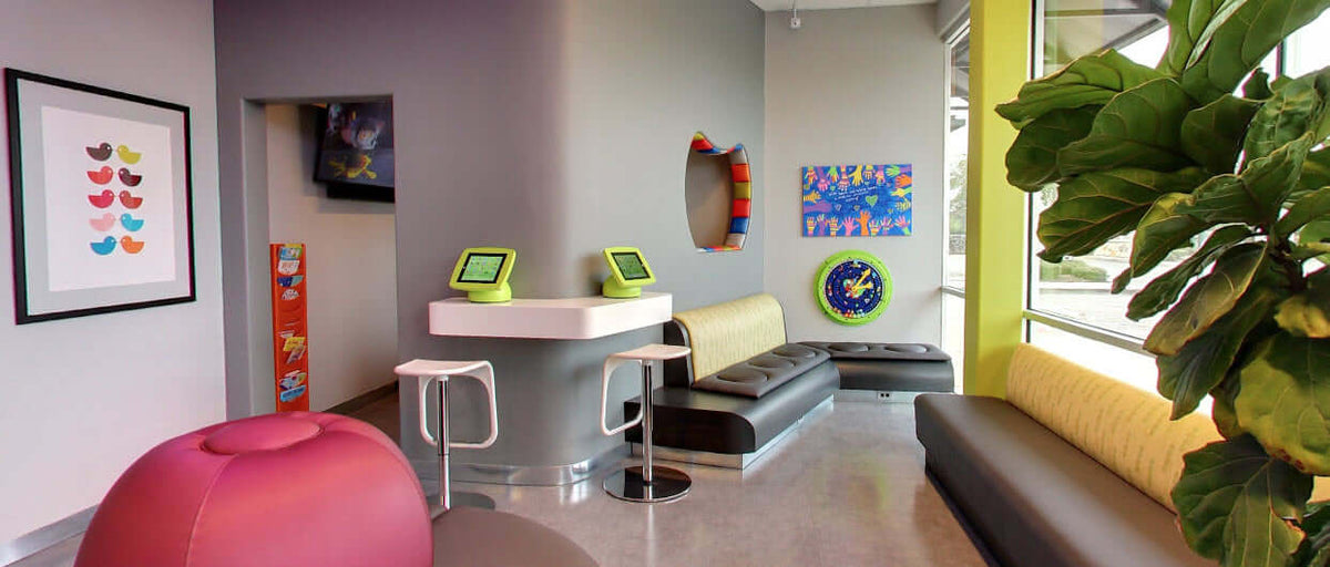 Pediatric dental office waiting area with two green Armodilo Sphere tablet kiosks on a counter, wall art, and a pink bean-bag chair