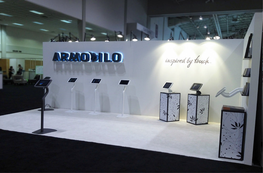 Armodilo trade show booth displaying white tablet floor stands, desktop stands on pedestals and a wall mount